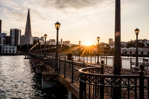 A sunset view from a pier with city buildings and a tall spire in the background, casting warm light over the water.