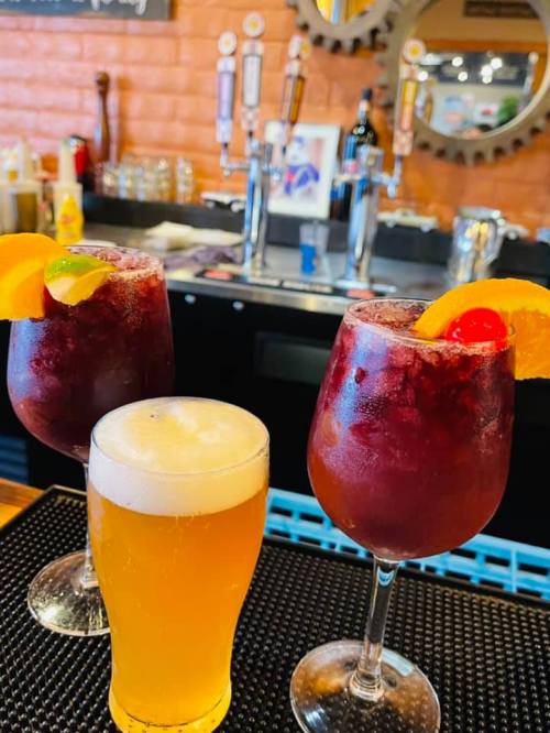 Three drinks on a bar: two large cocktails with fruit garnishes and a smaller glass of beer.