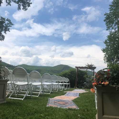 A scenic outdoor wedding setup with white chairs, a floral arch, and mountains in the background under a blue sky.