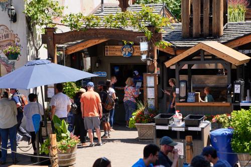 A bustling outdoor market scene with people gathered around food stalls and shaded areas, surrounded by greenery.