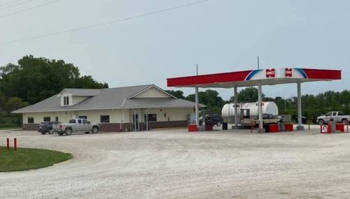 A gas station with a convenience store, surrounded by gravel, featuring several parked vehicles and a cloudy sky.