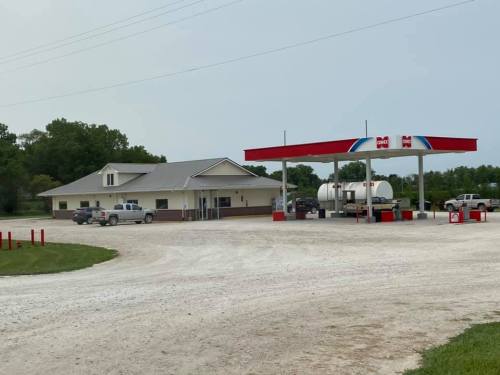 A gas station with a convenience store, surrounded by gravel and green trees, with several vehicles parked nearby.