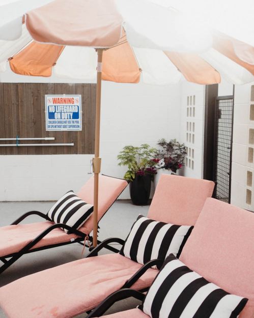 Two pink lounge chairs with striped cushions under an orange and white umbrella, near a "No Lifeguard" sign.