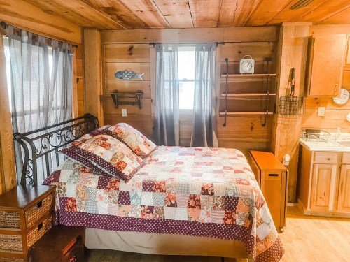 Cozy wooden bedroom with a quilted bed, window, and small kitchen area, featuring rustic decor and warm lighting.