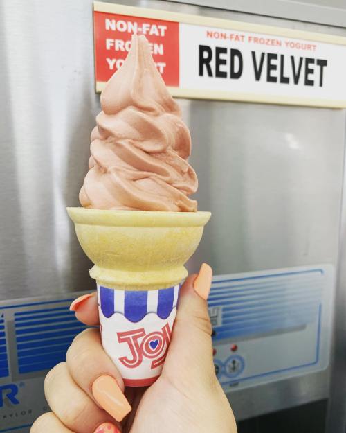 A hand holding a cone of red velvet frozen yogurt in front of a sign indicating the flavor.
