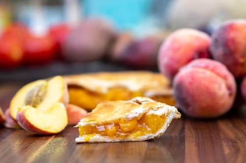 A slice of peach pie with fresh peaches in the background on a wooden surface.
