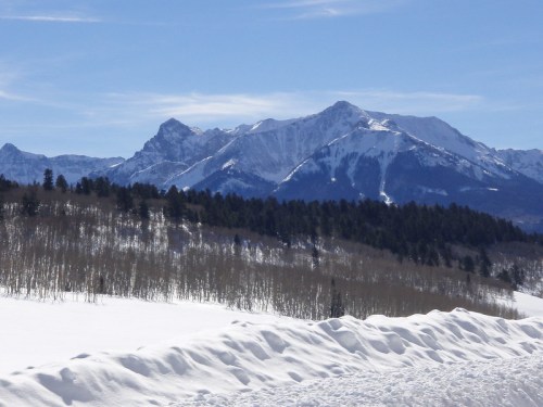 Snow-covered mountains rise against a clear blue sky, with a foreground of snowdrifts and sparse trees.
