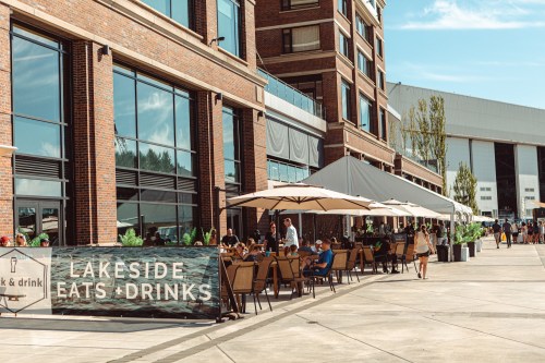 Outdoor dining area at Lakeside Eats & Drinks, with patrons enjoying meals under umbrellas by a modern building.