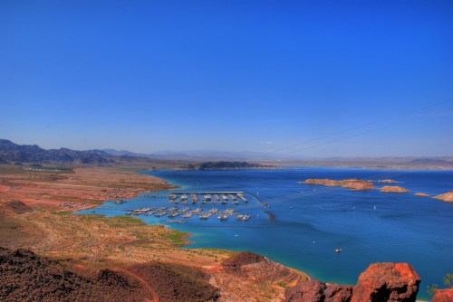 A scenic view of a blue lake surrounded by mountains, with boats docked at a marina under a clear blue sky.