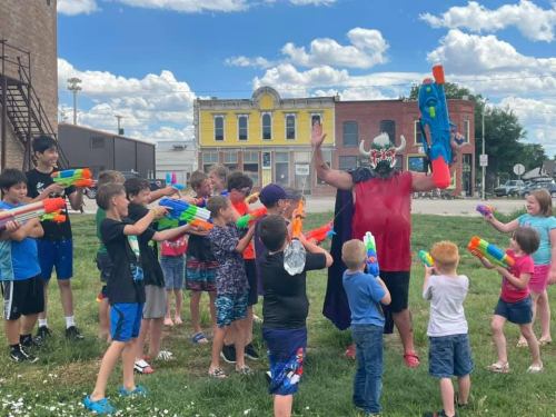 A group of children and teens play with water guns, while a person in a mask leads the fun in a grassy area.