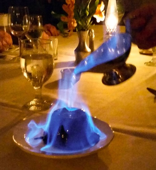 A dessert being flambéed at a table, with blue flames rising from a dome-shaped dish. Soft lighting in the background.