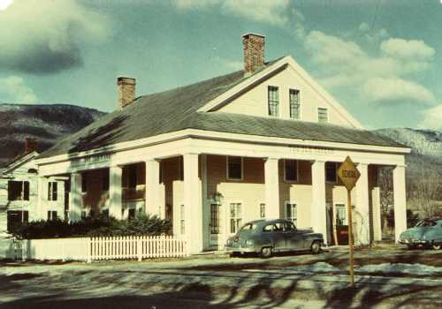 Historic building with columns, a school sign, and vintage cars parked outside, set against a mountainous backdrop.