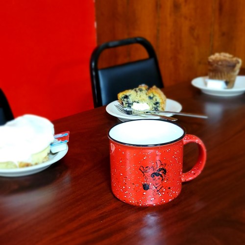 A red mug with a cartoon design sits on a table with blueberry muffins and a dessert plate in the background.