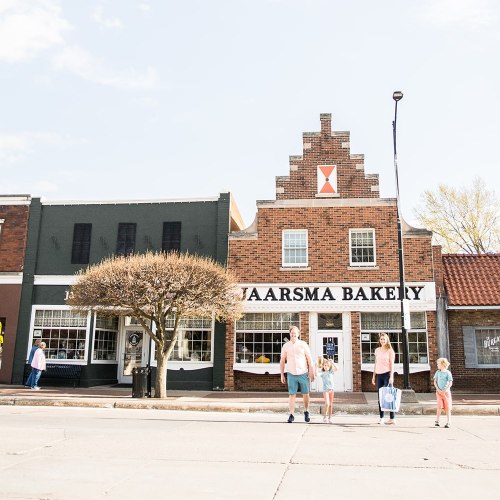 A family walks in front of Jaarsma Bakery, a charming brick building with a decorative gable, on a sunny day.