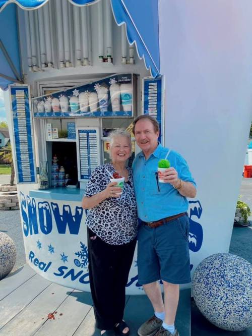 A smiling couple holds colorful snow cones in front of a snow cone stand, enjoying a sunny day.