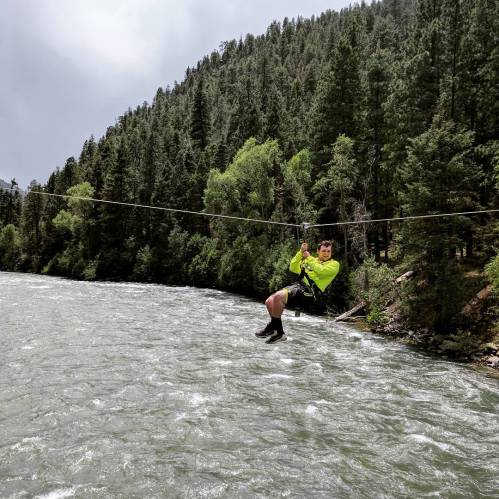A person in a bright yellow shirt zip-lining over a river surrounded by lush green trees and mountains.