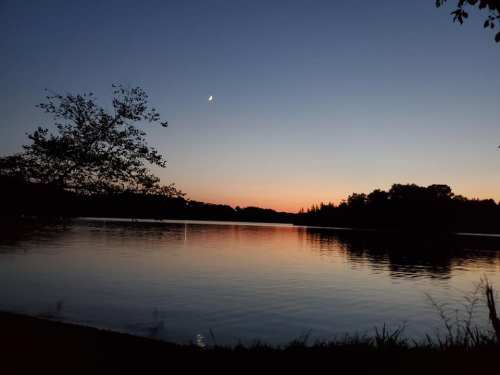 A serene lake at sunset, with a crescent moon and silhouettes of trees against a colorful sky.