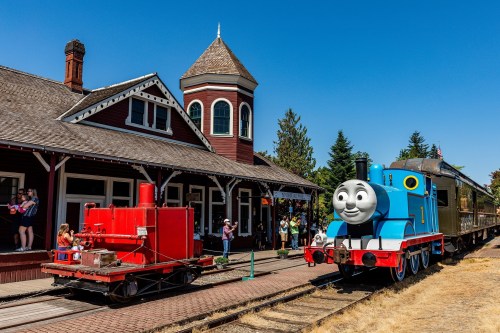 A blue cartoon train with a smiling face is parked at a station, alongside a red engine and visitors enjoying the scene.
