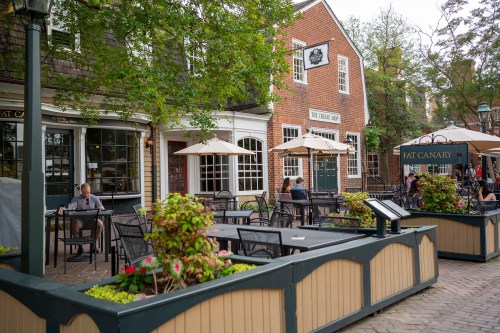 Outdoor dining area with tables, umbrellas, and greenery, featuring brick buildings in a charming neighborhood.