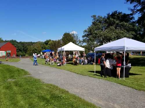 A sunny outdoor market scene with tents, vendors, and visitors on a grassy area. Trees and a red barn in the background.