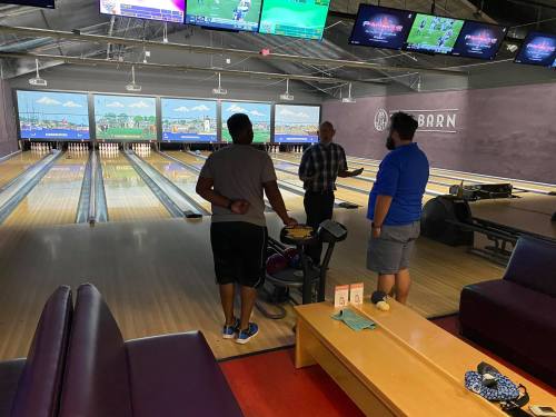 Three people converse near bowling lanes, with screens displaying scores in the background. A cozy lounge area is visible.