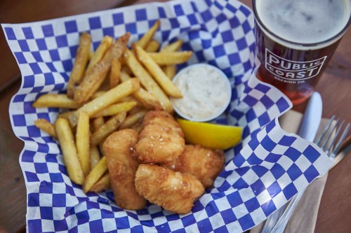A basket of golden fried fish bites with fries, a lemon wedge, and a dipping sauce, alongside a pint of beer.