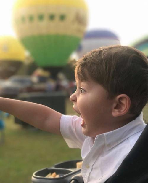 A young boy excitedly points at colorful hot air balloons in the background, with a joyful expression on his face.