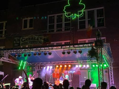 A lively band performs on stage at the Greeley Irish Festival, with colorful lights and a shamrock sign above.