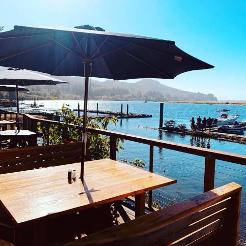 A sunny waterfront view from a wooden deck with tables, umbrellas, and boats docked in the background.