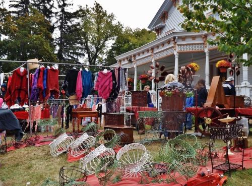 A vibrant outdoor market featuring colorful clothing and vintage items displayed on tables and racks near a historic house.