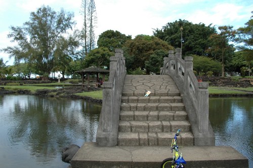 A stone bridge over a calm pond, surrounded by lush greenery and trees under a clear blue sky.