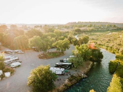 Aerial view of an RV park by a winding river, surrounded by trees and grassy areas under a warm sunset.