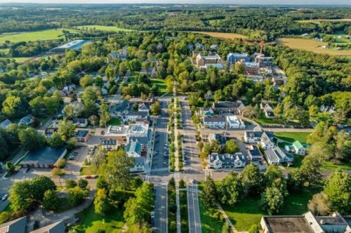 Aerial view of a small town with tree-lined streets, residential homes, and green fields in the background.