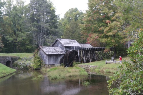 A rustic wooden mill with a waterwheel beside a calm pond, surrounded by trees in autumn colors.