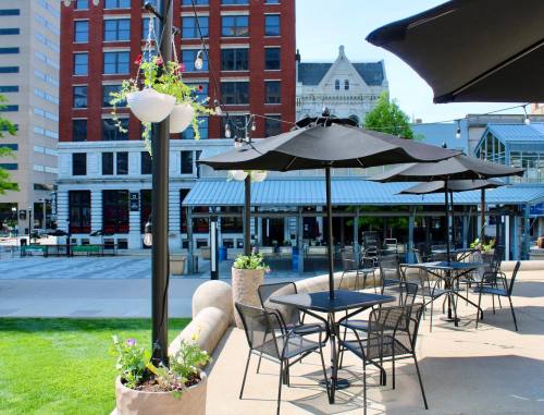 Outdoor dining area with black umbrellas and tables, surrounded by greenery and buildings in a city setting.
