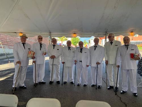 A group of eight men in white suits and black ties stand under a tent, holding canes and boxes, smiling at the camera.