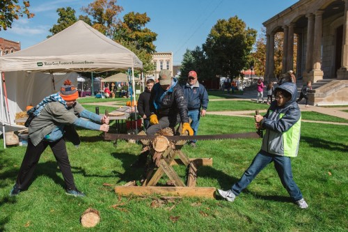 Two people saw a log with a hand saw while others watch at an outdoor event, with tents and trees in the background.
