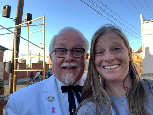 A man in a white suit and bow tie smiles with a woman in a casual shirt, both posing for a selfie outdoors.