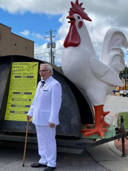 A man in a white suit stands next to a giant rooster statue and a large frying skillet display.