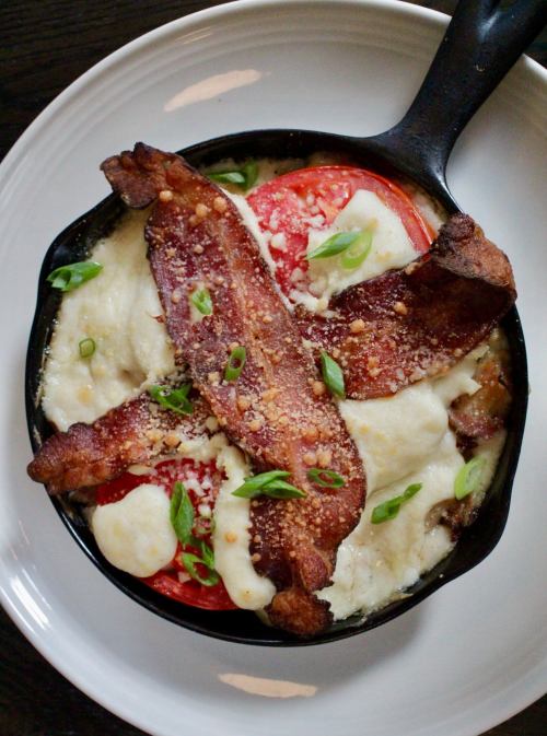 A cast-iron skillet filled with creamy dish topped with bacon, tomatoes, and green onions.