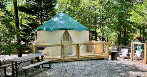 A yurt with a green roof sits in a wooded area, surrounded by a wooden deck, picnic table, and outdoor seating.