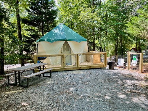 A yurt with a green roof sits in a wooded area, featuring a wooden deck and picnic table nearby.