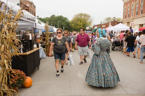 A bustling street market with vendors, shoppers, and a woman in a vintage dress walking among the crowd.