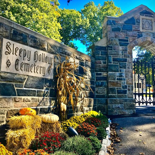 Entrance to Sleepy Hollow Cemetery, featuring stone walls, autumn decorations, pumpkins, and colorful flowers.
