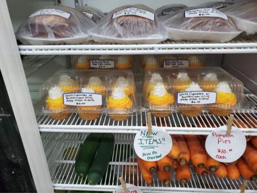 A fridge display with various desserts, including pumpkin pies and cupcakes topped with frosting, alongside fresh vegetables.
