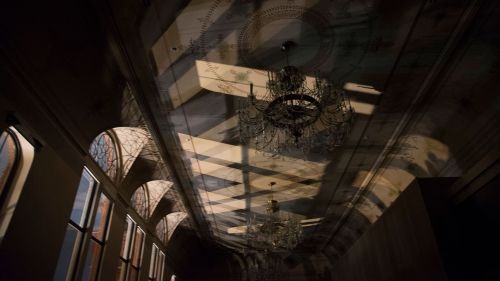 A dimly lit hallway with ornate chandeliers and intricate shadows cast on the walls and ceiling.