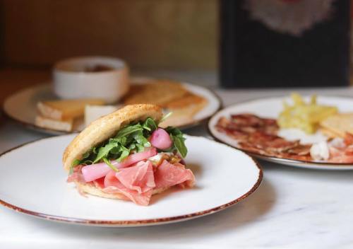 A sandwich with ham, arugula, and pickled vegetables on a plate, with toast and a charcuterie board in the background.