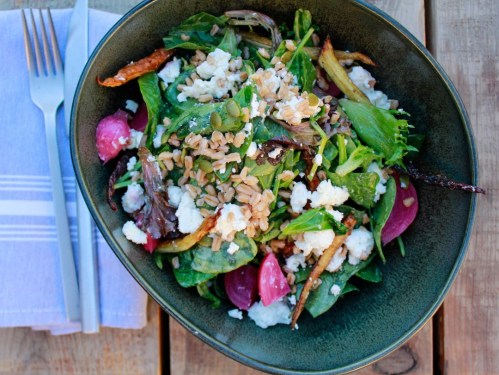 A vibrant salad with greens, roasted vegetables, goat cheese, and seeds in a dark bowl on a wooden table.