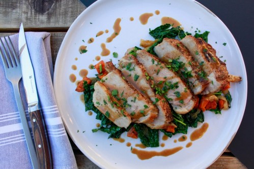 A plate of sliced pork chop on a bed of sautéed greens and tomatoes, drizzled with sauce, next to a fork and knife.
