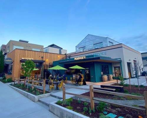 A modern market and kitchen with outdoor seating, surrounded by greenery and a clear blue sky.